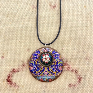 May include: A circular pendant necklace with a black cord. The pendant features a blue, gold, and red floral design with a smaller floral centerpiece. The necklace is likely handmade and suitable for everyday wear.
