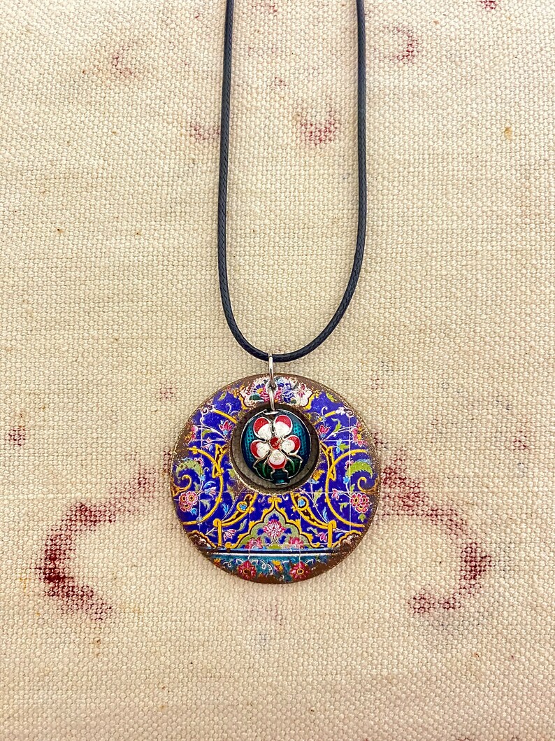 May include: A circular pendant necklace with a black cord. The pendant features a blue, gold, and red floral design with a smaller floral centerpiece. The necklace is likely handmade and suitable for everyday wear.