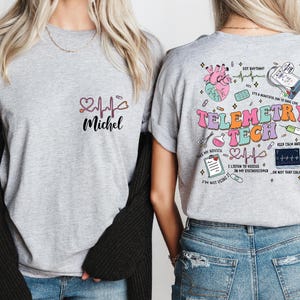 May include: A light gray t-shirt with the name "Michel" and a heart and EKG design on the front. The back of the shirt features the words "Telemetry Tech" with medical-themed graphics, including a heart, monitor, and stethoscope.