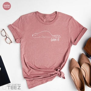 May include: A dusty rose t-shirt with a white outline of a beaver and the words "DAM IT" printed below. The shirt is knotted at the bottom right. A pair of glasses and shoes are visible in the background.