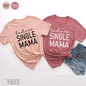 May include: Two short-sleeved t-shirts, one in heather peach and the other in heather mauve, each with the words "badass SINGLE MAMA" printed on the front in a bold font. The peach shirt has black text, while the mauve shirt has white text.