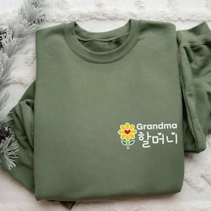 May include: An olive green sweatshirt with a crew neck. The front features a graphic of a yellow flower with a red heart center and the word "Grandma" in English and Korean script. The sweatshirt is folded.