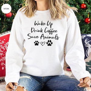 May include: White sweatshirt with the text "Wake Up Drink Coffee Save Animals" in black script. The design includes paw prints and a heart. The sweatshirt is a long-sleeved crew neck.