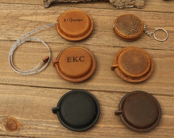 Peut inclure: Plusieurs mètres rubans en cuir de différentes nuances de marron et de noir. Certains sont gaufrés de motifs floraux ou personnalisés avec des initiales et le texte "#1 Grandpa". Les mètres rubans sont sur une surface en bois.