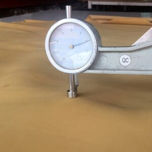 May include: A close-up of a dial gauge measuring the thickness of a piece of tan leather. The gauge reads 5.5 mm. The gauge is labeled "QC".