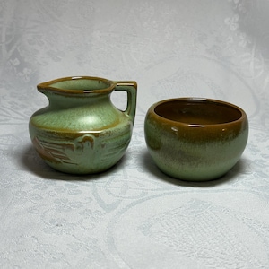 May include: Two small, handcrafted ceramic pieces. One is a small pitcher with a handle, and the other is a round bowl. Both are glazed in a green color with brown accents, suggesting a rustic or vintage aesthetic.