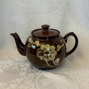 May include: A brown ceramic teapot with a lid and handle. The teapot features a floral design in gold and cream. The teapot is on a white surface with a floral pattern.