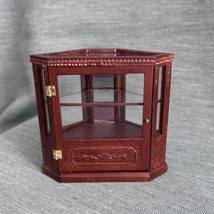 May include: A miniature wooden display cabinet with a hexagonal shape. The cabinet has glass panels, interior shelves, and a small door. The wood is a dark brown color, and the cabinet has decorative carvings.