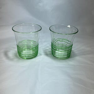 May include: Two clear glass tumblers with a green, wavy, horizontal design around the base. The glasses have a slightly flared shape and are set against a white background. The green design adds a pop of color.