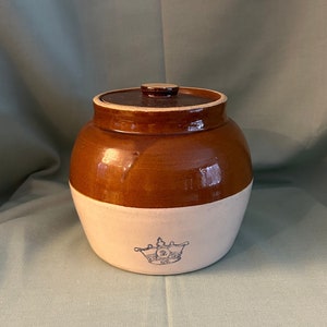 May include: A brown and white glazed ceramic crock with a lid. The crock has a crown design on the front.