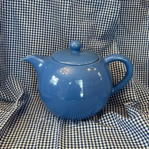 May include: A blue ceramic teapot with a lid and a handle. The teapot is a solid, light blue color and has a rounded shape. The background is a blue and white checkered pattern.