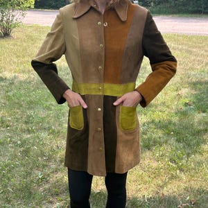 May include: A long-sleeved suede coat featuring a vertical patchwork design in various shades of brown, tan, and olive green. The coat includes a pointed collar, a front button closure with metallic buttons, and two olive green patch pockets at the waist.