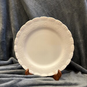 May include: A white ceramic plate with a scalloped edge and embossed floral design. The plate is displayed on a wooden stand against a gray background. The plate's diameter is approximately 10 inches.