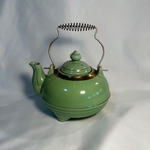 May include: A light green ceramic teapot with a gold rim and handle. The teapot has a rounded body, a lid with a small knob, and a spout. The handle is made of metal and has a coiled design. The teapot sits on three small feet.