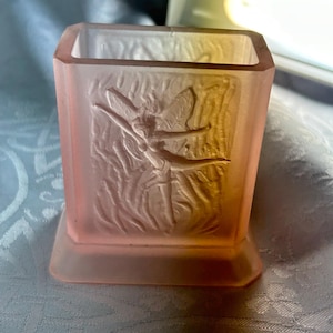 May include: A pink, rectangular glass container with a frosted finish. The front of the container features a raised relief of a fairy. The container has a square base and a rectangular opening at the top. The glass has a slight gradient of color.