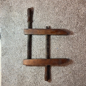 May include: A wooden clamp with two adjustable screws and wooden handles. The clamp is made of dark brown wood and has a rustic appearance.