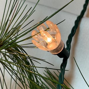 May include: A close-up of a clear, plastic Christmas light bulb with a warm, white glow. The bulb is attached to a green wire cord.