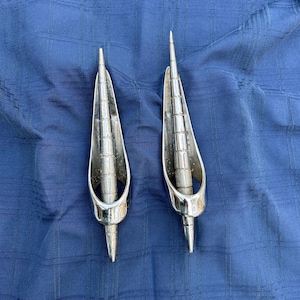 May include: Two silver-toned metal car hood ornaments shaped like a pointed arrow with a ring at the base.