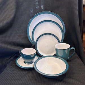 May include: A dinnerware set featuring white centers and dark teal rims. The collection includes three stacked round plates of varying sizes, a bowl, a mug, and a teacup resting on a matching saucer. Each piece has a smooth, glossy finish.