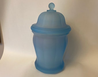 May include: A frosted blue glass jar with a lid and a knob on top. The jar has a rounded base and a slightly flared top.