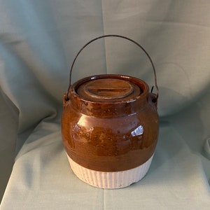 May include: A brown and white glazed ceramic crock with a wire handle and a lid. The crock has a white band around the middle.