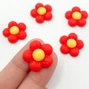 Puede incluir: Cinco adornos de color rojo en forma de flor con centros amarillos. Las flores están hechas de un material suave y flexible.