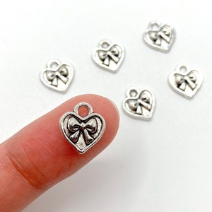 May include: Silver-colored heart-shaped charms with a bow design. The charms have a loop at the top for attaching to jewelry. One charm is held by a finger, with several others scattered on a white surface. These charms are suitable for DIY jewelry making.