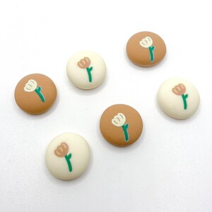 May include: Six small round resin buttons with a floral design. The buttons are in two colors: brown and white. Each button features a stylized flower with a white center and brown petals. The flowers have green stems.