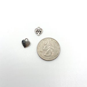 May include: Two small silver heart charms and a quarter coin are displayed on a white surface. One heart charm is solid, the other has a bow design. The quarter coin is silver and shows a profile of George Washington.