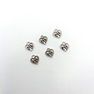 May include: Six small, silver-colored heart charms with a bow design. Each charm has a loop at the top for attaching to jewelry. The charms are scattered on a white surface.