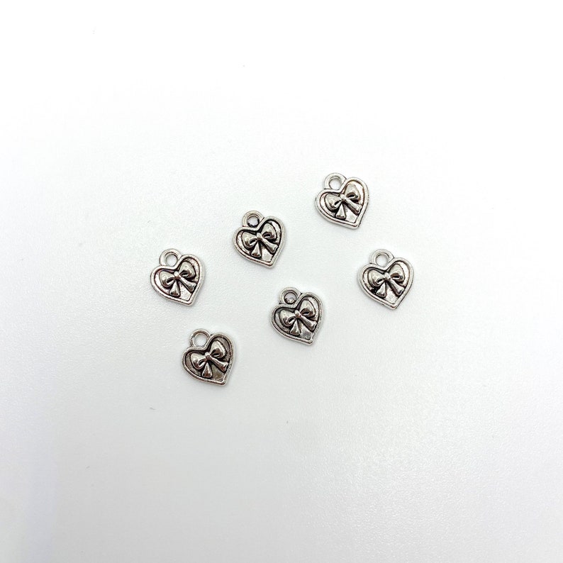 May include: Six small, silver-colored heart charms with a bow design. Each charm has a loop at the top for attaching to jewelry. The charms are scattered on a white surface.