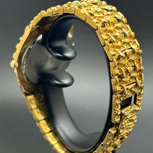 May include: A gold-tone wristwatch featuring a highly textured, nugget-style link bracelet. The watch case is round with a gold-tone finish, and the band consists of interconnected, irregular metallic segments that create a rugged, raised surface appearance.