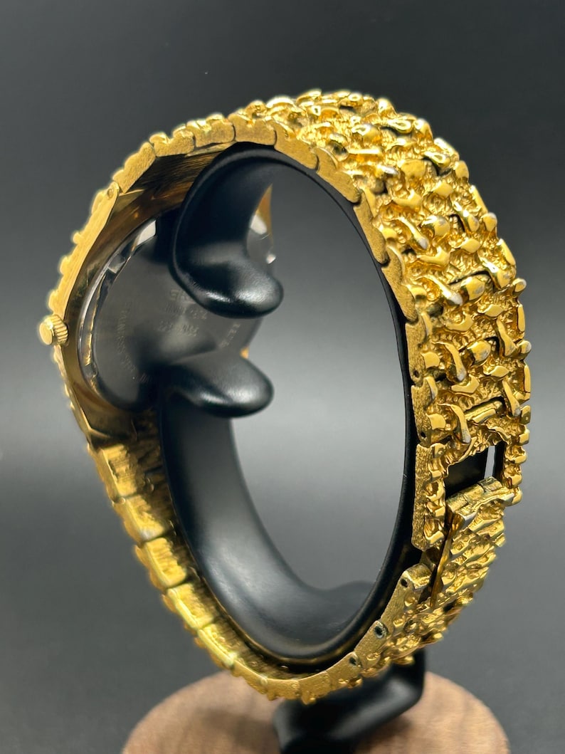 May include: A gold-tone wristwatch featuring a highly textured, nugget-style link bracelet. The watch case is round with a gold-tone finish, and the band consists of interconnected, irregular metallic segments that create a rugged, raised surface appearance.