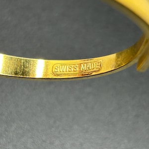 Puede incluir: Primer plano de una pulsera dorada con un acento rojo. La pulsera tiene las palabras "SWISS MADE" estampadas en el interior. El fondo es oscuro.