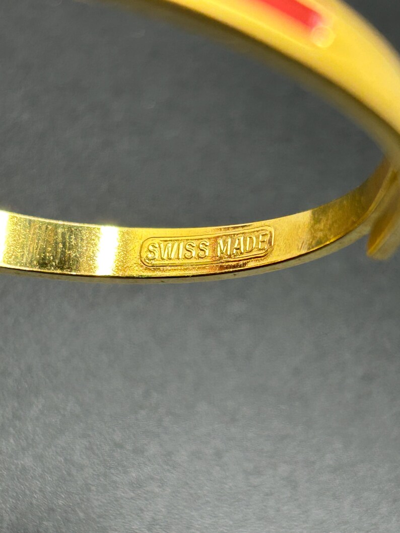 Puede incluir: Primer plano de una pulsera dorada con un acento rojo. La pulsera tiene las palabras "SWISS MADE" estampadas en el interior. El fondo es oscuro.