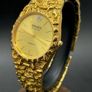 May include: A gold-tone Gruen Diamond quartz watch featuring a textured, nugget-style link bracelet and a round gold dial. The watch face displays a single diamond marker at the twelve o'clock position, thin gold hands, and the text Gruen Diamond Quartz.