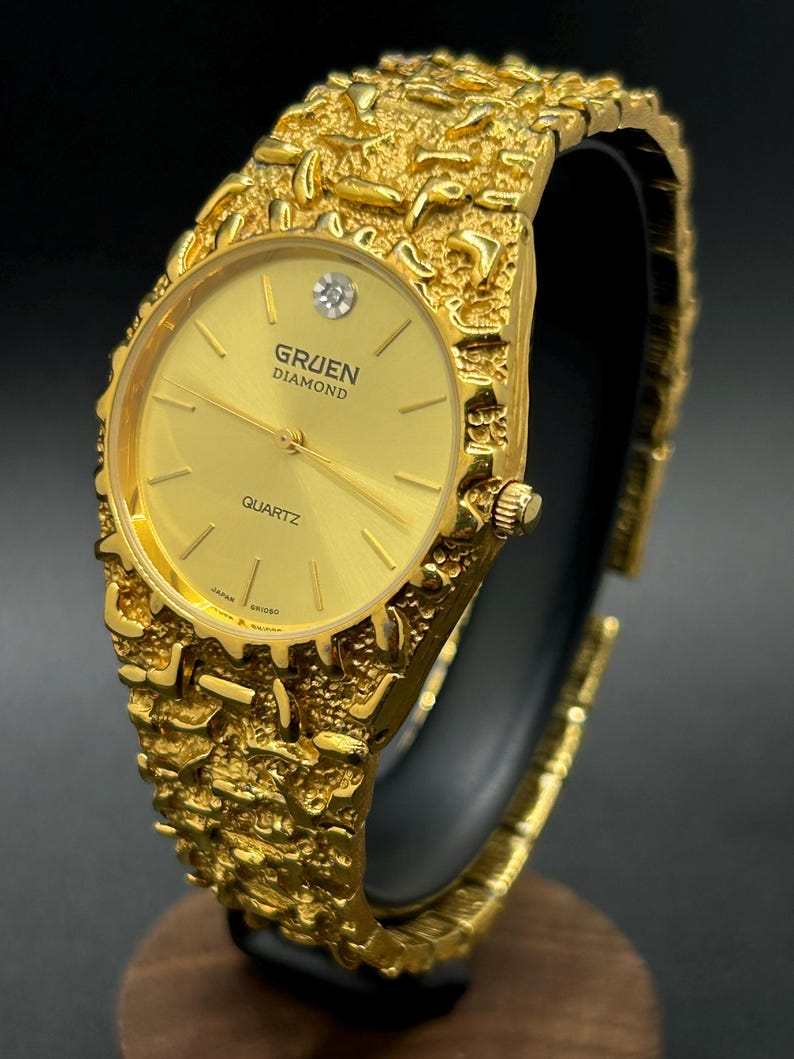 May include: A gold-tone Gruen Diamond quartz watch featuring a textured, nugget-style link bracelet and a round gold dial. The watch face displays a single diamond marker at the twelve o'clock position, thin gold hands, and the text Gruen Diamond Quartz.
