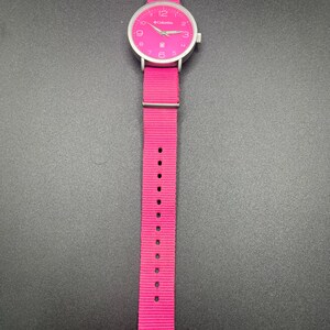May include: A bright pink Columbia watch with a silver-colored case and a matching pink nylon strap. The watch face is pink with white numerals and hands. The watch is displayed against a dark background.