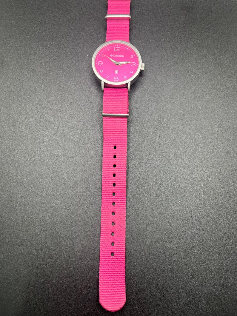 May include: A bright pink Columbia watch with a silver-colored case and a matching pink nylon strap. The watch face is pink with white numerals and hands. The watch is displayed against a dark background.