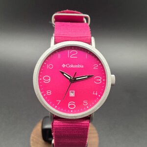 May include: A vibrant pink Columbia watch with a silver-tone case and a matching pink fabric band. The watch face displays the Columbia logo, numerals, and a date window. The watch is displayed on a wooden stand.