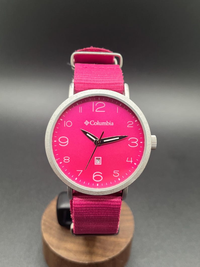 May include: A vibrant pink Columbia watch with a silver-tone case and a matching pink fabric band. The watch face displays the Columbia logo, numerals, and a date window. The watch is displayed on a wooden stand.