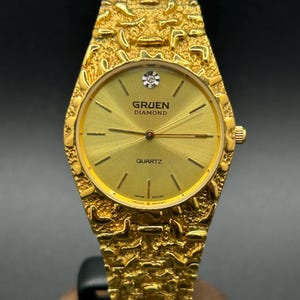May include: A gold-tone Gruen Diamond quartz watch with a textured, nugget-style link bracelet and case. The round dial is gold with thin hour markers, a single diamond at the twelve o'clock position, and the text Gruen Diamond Quartz.