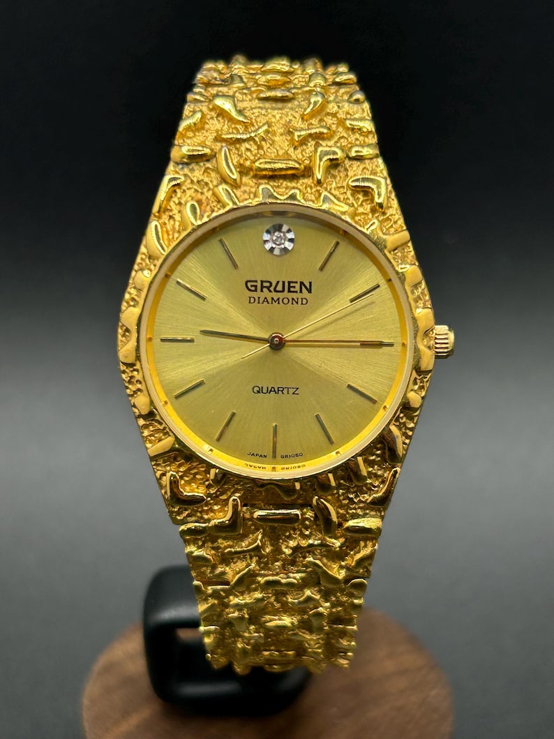 May include: A gold-tone Gruen Diamond quartz watch with a textured, nugget-style link bracelet and case. The round dial is gold with thin hour markers, a single diamond at the twelve o'clock position, and the text Gruen Diamond Quartz.