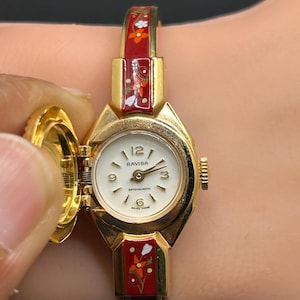 Puede incluir: Un reloj de pulsera Ravisa vintage en tono dorado con esfera blanca y n&uacute;meros negros. La correa del reloj es roja con dise&ntilde;os florales. La esfera del reloj est&aacute; abierta, revelando la hora. El reloj es de fabricaci&oacute;n suiza.