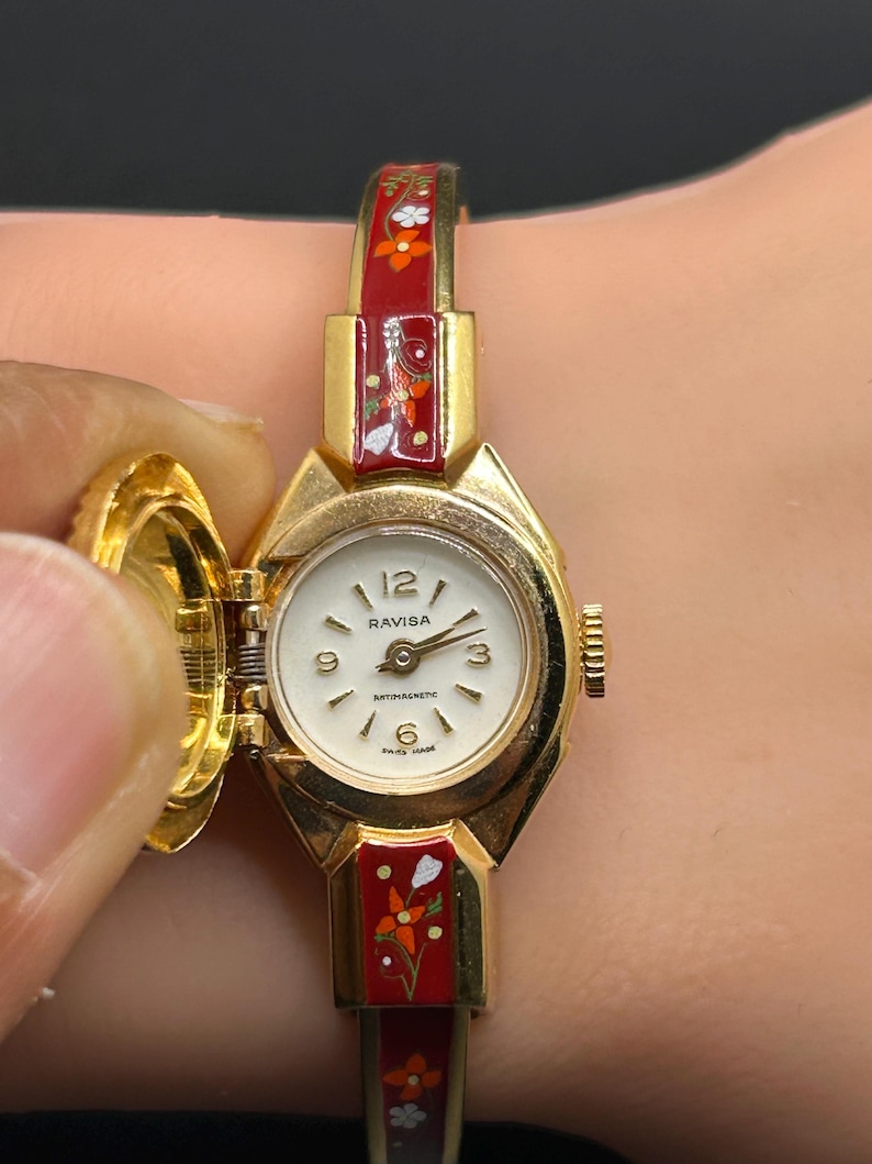 Puede incluir: Un reloj de pulsera Ravisa vintage en tono dorado con esfera blanca y n&uacute;meros negros. La correa del reloj es roja con dise&ntilde;os florales. La esfera del reloj est&aacute; abierta, revelando la hora. El reloj es de fabricaci&oacute;n suiza.