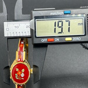 Puede incluir: Un reloj de pulsera vintage dorado con esfera y correa de esmalte rojo, adornado con dise&ntilde;os florales. La esfera del reloj es redonda y la correa mide aproximadamente 19,7 mm de ancho. El reloj se muestra sobre un fondo oscuro.