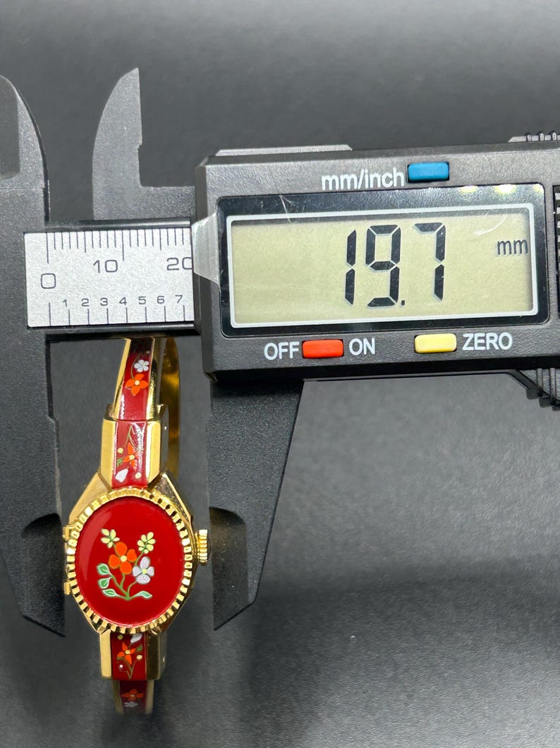 Puede incluir: Un reloj de pulsera vintage dorado con esfera y correa de esmalte rojo, adornado con dise&ntilde;os florales. La esfera del reloj es redonda y la correa mide aproximadamente 19,7 mm de ancho. El reloj se muestra sobre un fondo oscuro.