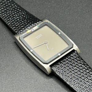 K&ouml;nnte beinhalten: Eine Vintage Ricoh Quartz-Uhr mit quadratischem, taupefarbenem Zifferblatt und silberfarbenem Geh&auml;use. Die Uhr hat ein schwarzes, strukturiertes Lederarmband. Das Zifferblatt zeigt den Markennamen und das Wort "QUARTZ".