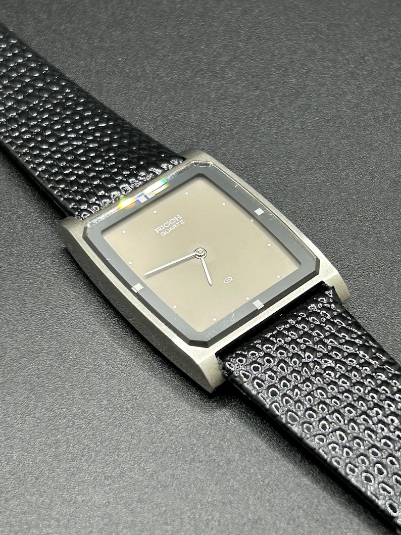 K&ouml;nnte beinhalten: Eine Vintage Ricoh Quartz-Uhr mit quadratischem, taupefarbenem Zifferblatt und silberfarbenem Geh&auml;use. Die Uhr hat ein schwarzes, strukturiertes Lederarmband. Das Zifferblatt zeigt den Markennamen und das Wort "QUARTZ".