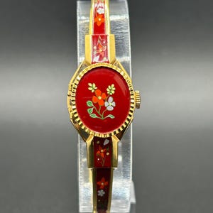 Puede incluir: Un reloj vintage dorado con esfera y correa rojas. La esfera del reloj presenta un dise&ntilde;o floral con detalles en naranja, blanco y verde. La correa tambi&eacute;n es roja con motivos florales. El reloj se exhibe sobre un soporte transparente.
